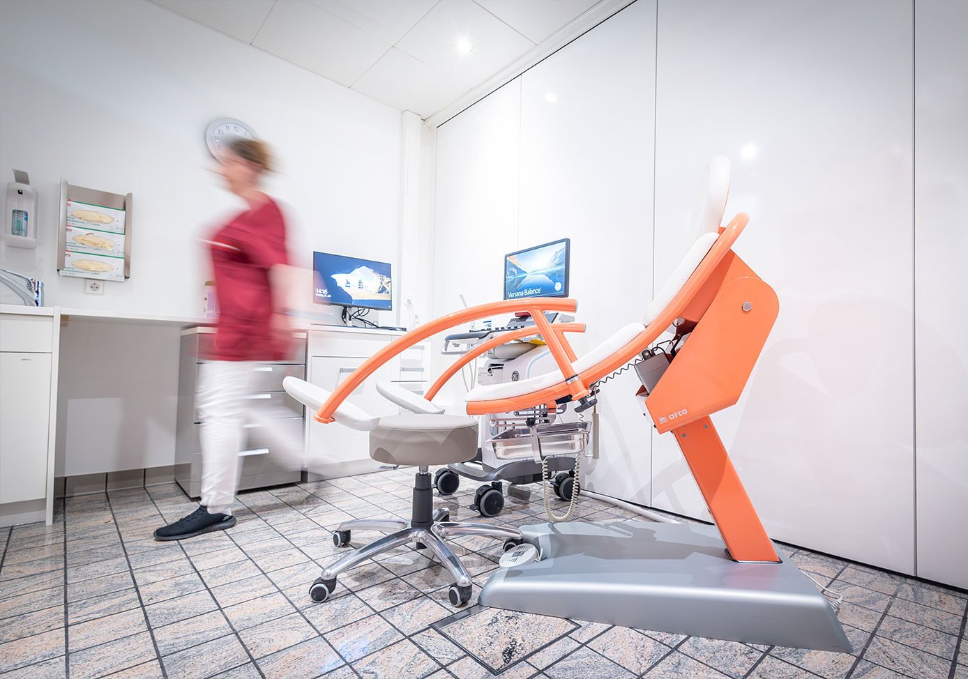 Behandlungsraum von Next Fertility Ulm mit einem gynäkologischen Behandlungsstuhl, zwei Monitoren und einer Mitarbeiterin.